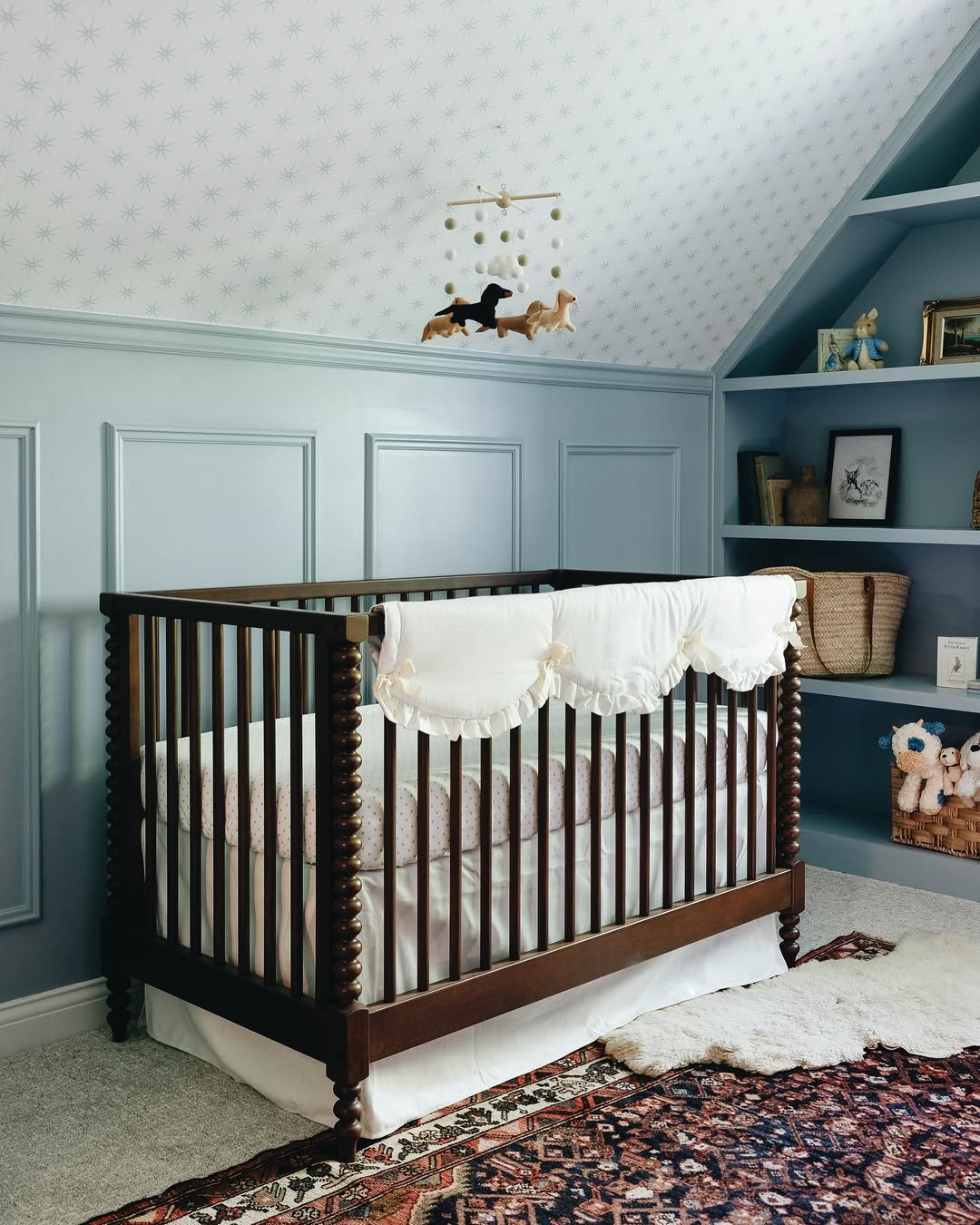 A dark wooden crib with white bedding stands against a blue paneled wall under a sloped ceiling with star-patterned wallpaper—an inspiring space for first-time moms seeking chic nursery ideas. Shelves with toys and baskets are on the right.