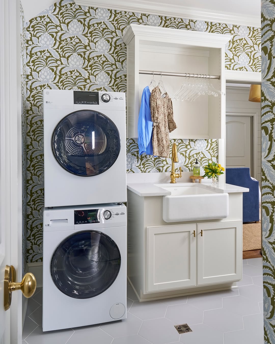 A compact stacked laundry room with a washer and dryer, a white sink with gold faucet, open shelving, hanging clothes, and floral wallpaper creates an organized yet stylish space.