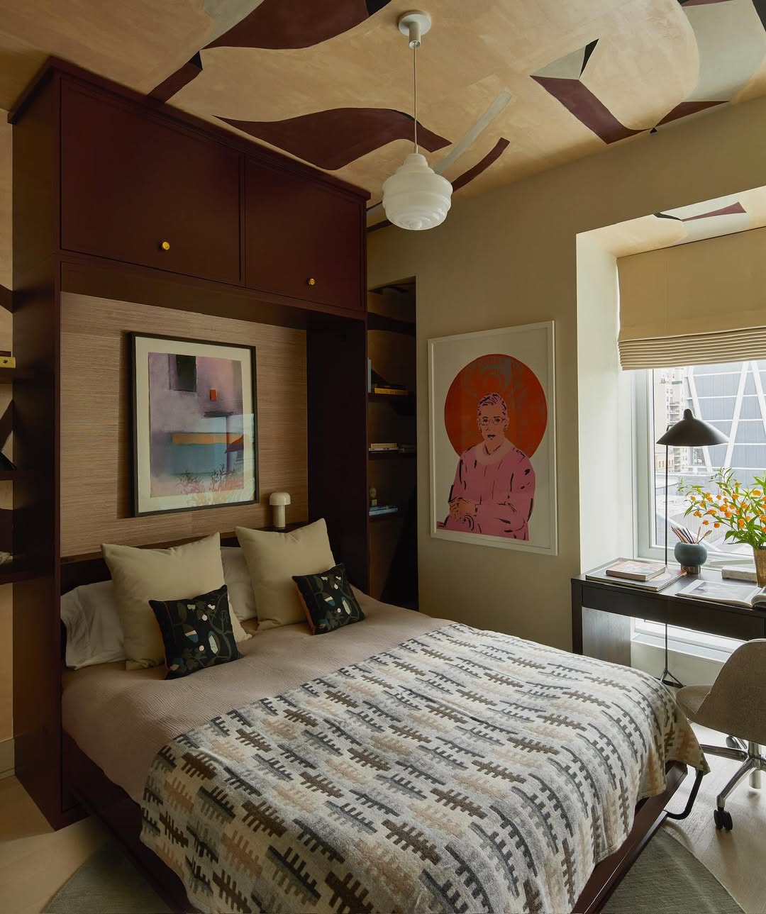 A modern bedroom with a patterned bedspread, abstract wall art, built-in shelves, a desk by the window, and a ceiling fan. Discover inspiring Murphy Bed Ideas that maximize space as natural light enters through a large window.
