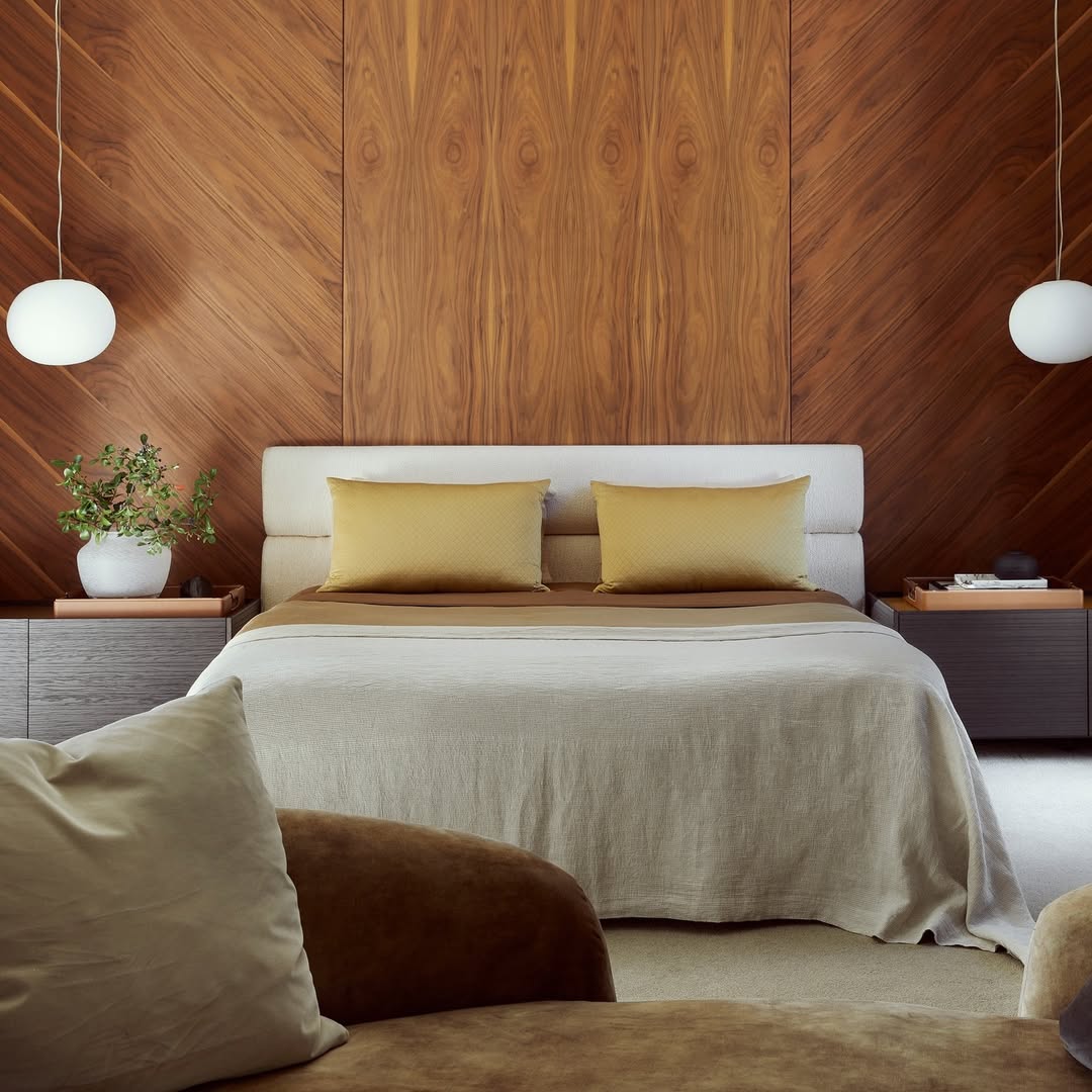 A modern bedroom design with a beige bed, two yellow pillows, wooden accent wall, round pendant lights, side tables, and a potted plant. A brown chair with a white cushion adds to the stylish bedroom decor in the foreground.