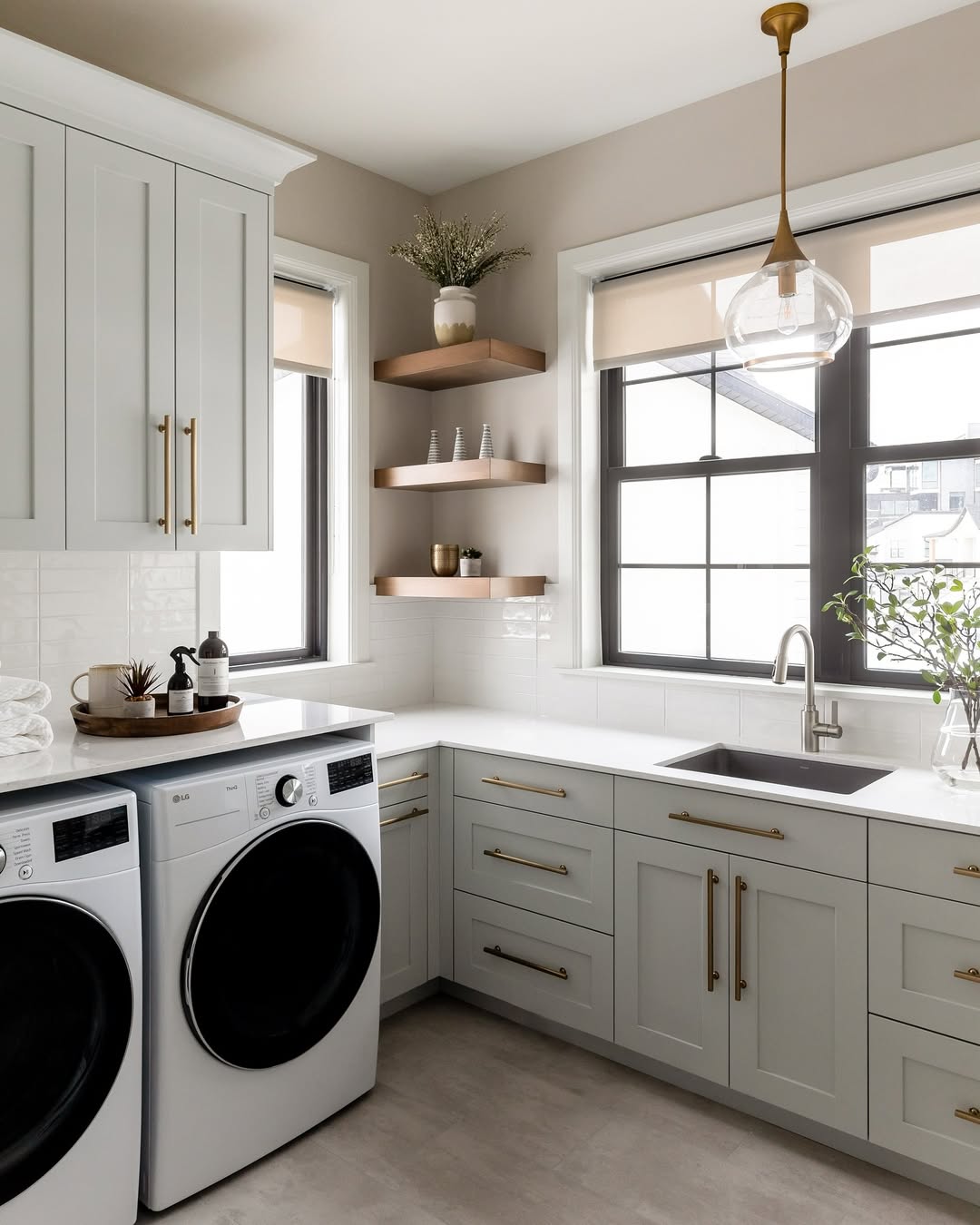Modern laundry room with stylish laundry room cabinets, white countertops, front-load washer and dryer, gold hardware, floating shelves, and large windows. Discover inspiring laundry room cabinet ideas for a fresh and functional space.