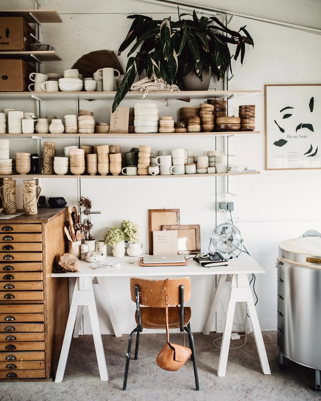 A pottery workspace doubles as a cozy home office workshop, with shelves displaying ceramic pieces and supplies above a desk featuring a laptop, dried flowers, and a brown chair in front.