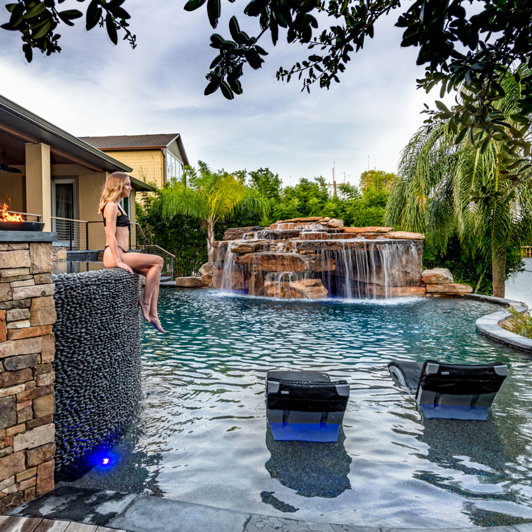 A woman in a swimsuit sits on the edge of a stone pool with a waterfall feature, lounge chairs in shallow water, and tropical landscaping—showcasing one of the coolest backyard pool ideas.