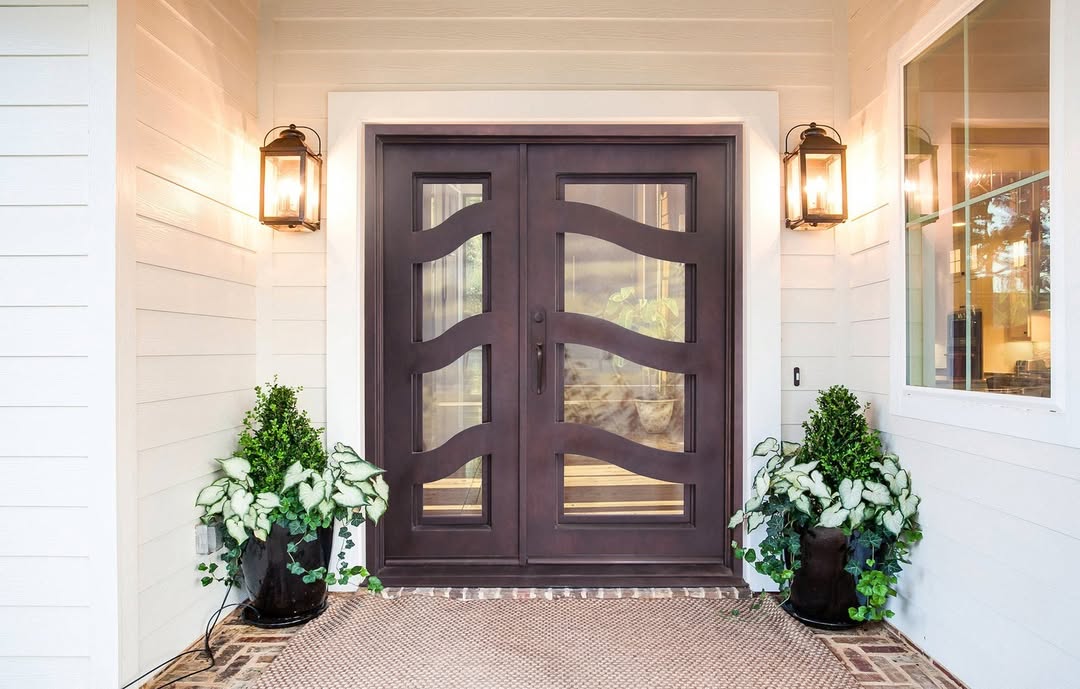 Double door entryway featuring wavy glass panels, flanked by potted plants and exterior wall lanterns against classic white siding—perfect for inspiring elegant double door designs and entryway ideas.