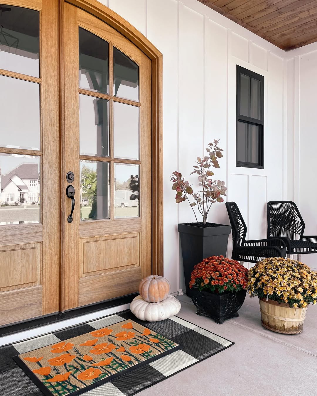 A charming double door entryway features wooden doors, a fall-themed doormat, a pumpkin, potted orange chrysanthemums, a black plant pot, and two black chairs beside a small table—perfect inspiration for cozy entryway ideas.