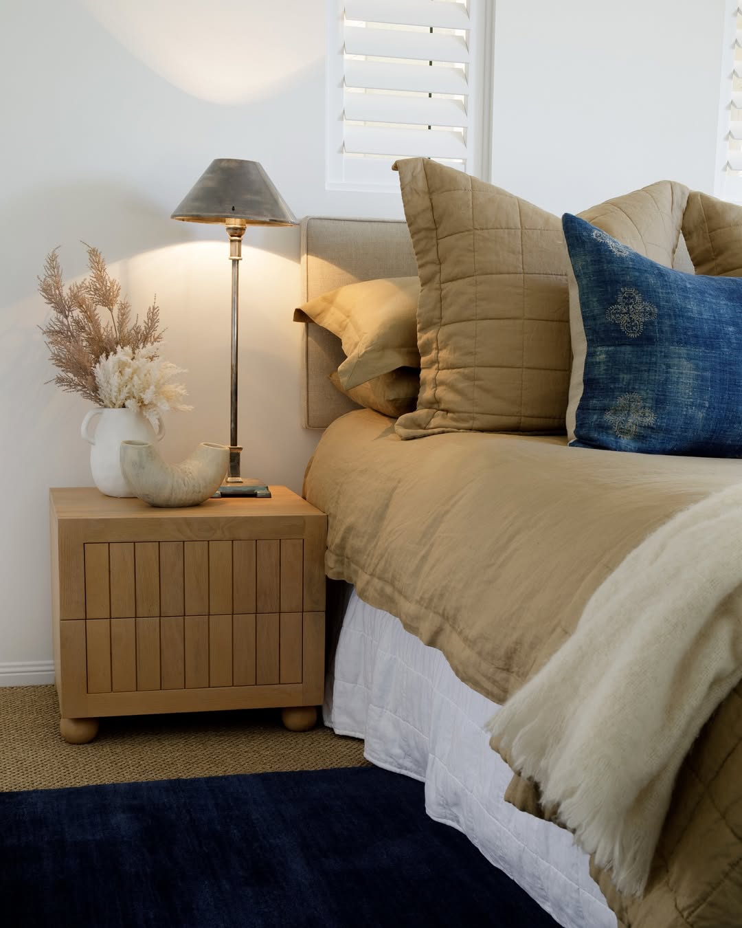 A neatly made bed with beige bedding and blue accent pillows sits next to a wooden nightstand holding a lamp, a ceramic vase with dried plants, and a decorative horn—perfect inspiration for 2025 bedroom color schemes for interior lovers.