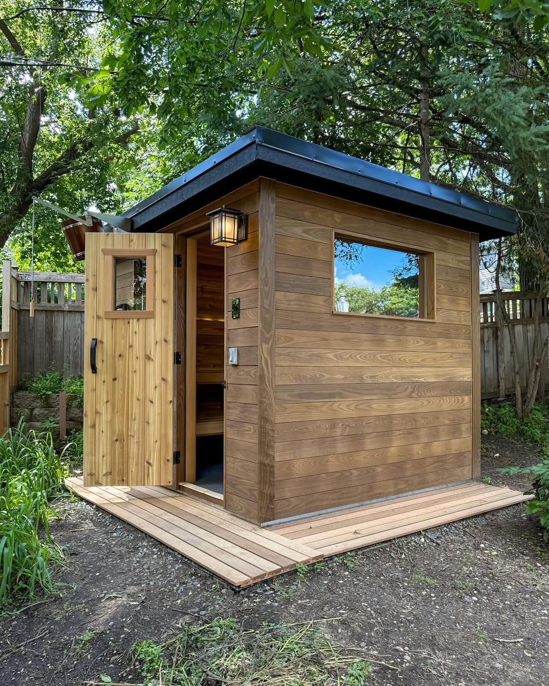 A small wooden outdoor sauna with an open door, a window, and a light fixture sits on a wood deck in a fenced backyard surrounded by trees, inspiring cozy outdoor sauna ideas.