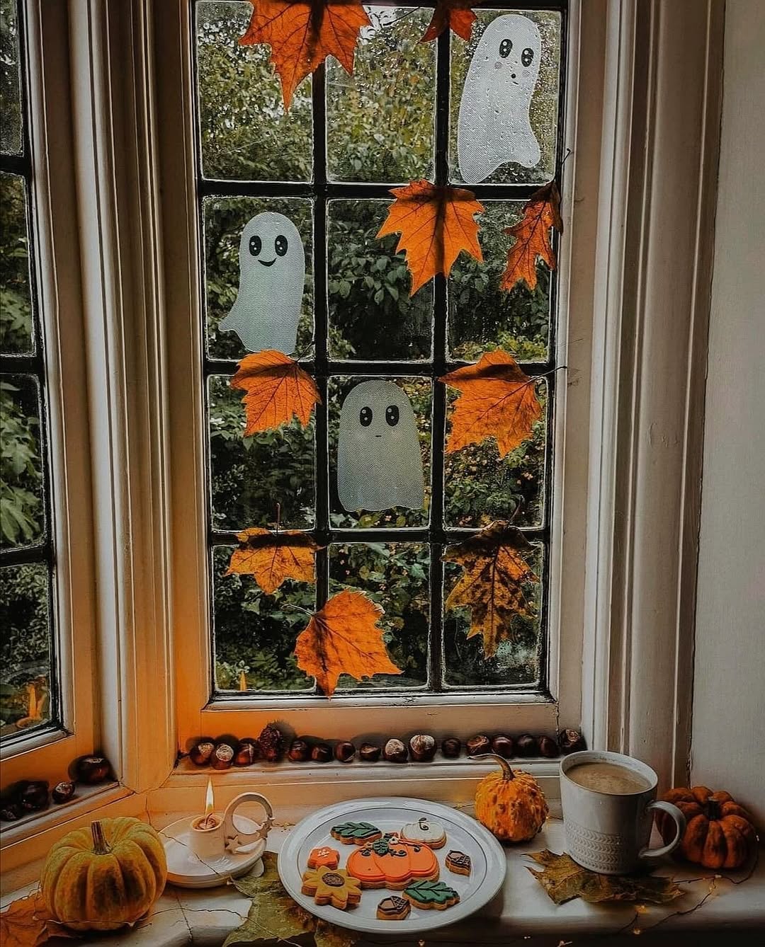 A window decorated with paper ghosts, autumn leaves, and other Halloween decorations overlooks trees; on the windowsill are cookies, a candle, a mug, a pumpkin, and chestnuts, creating a cozy autumn scene.