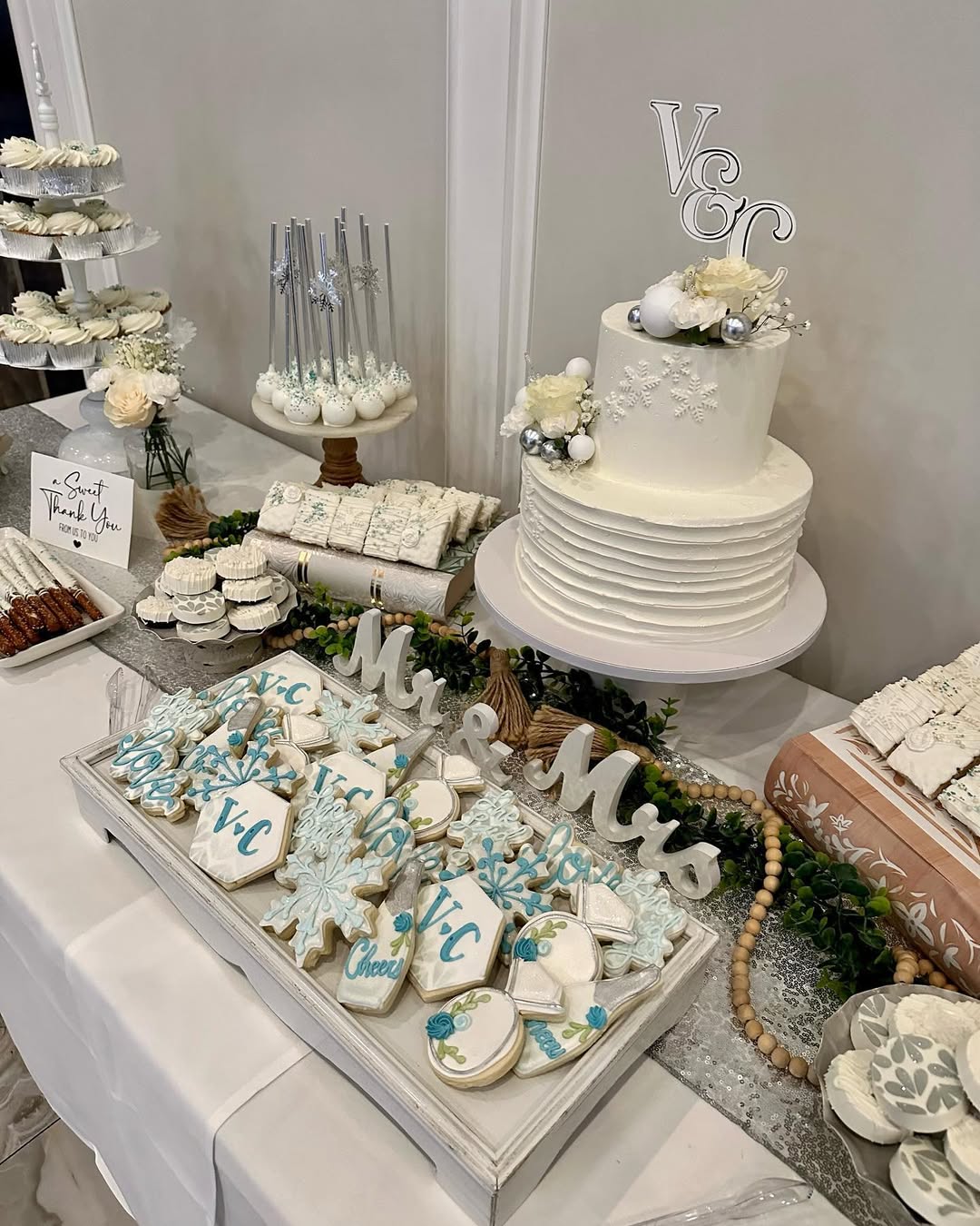 A wedding dessert table with a white tiered cake, blue and white decorated cookies, cake pops, and various pretty desserts arranged on trays and stands—perfect for those seeking elegant dessert table ideas.