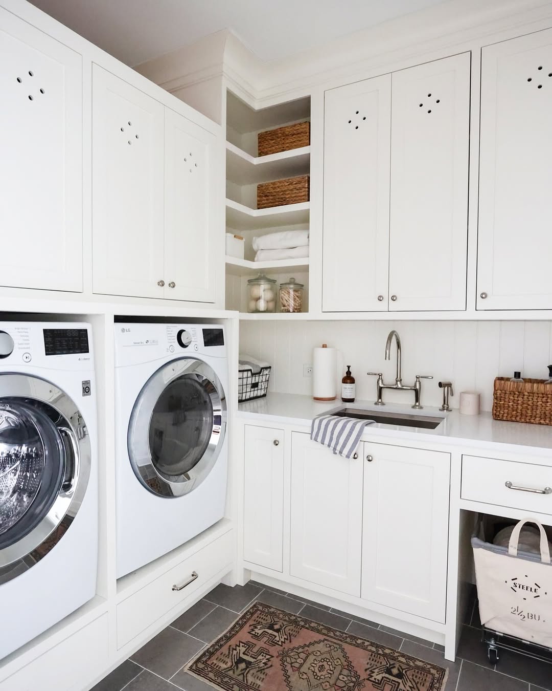 A laundry room with white laundry room cabinets, a front-loading washer and dryer, a built-in sink, open shelves with towels, and countertop accessories—perfect inspiration for stylish laundry room cabinet ideas.