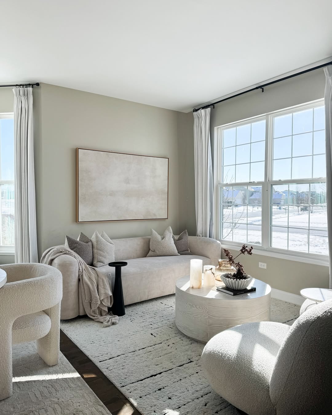 A bright, modern living room with neutral-toned furniture, large windows, light curtains, a round coffee table, and abstract wall art. Sunlight fills this expensive looking living room, perfect for those seeking modern living room ideas.