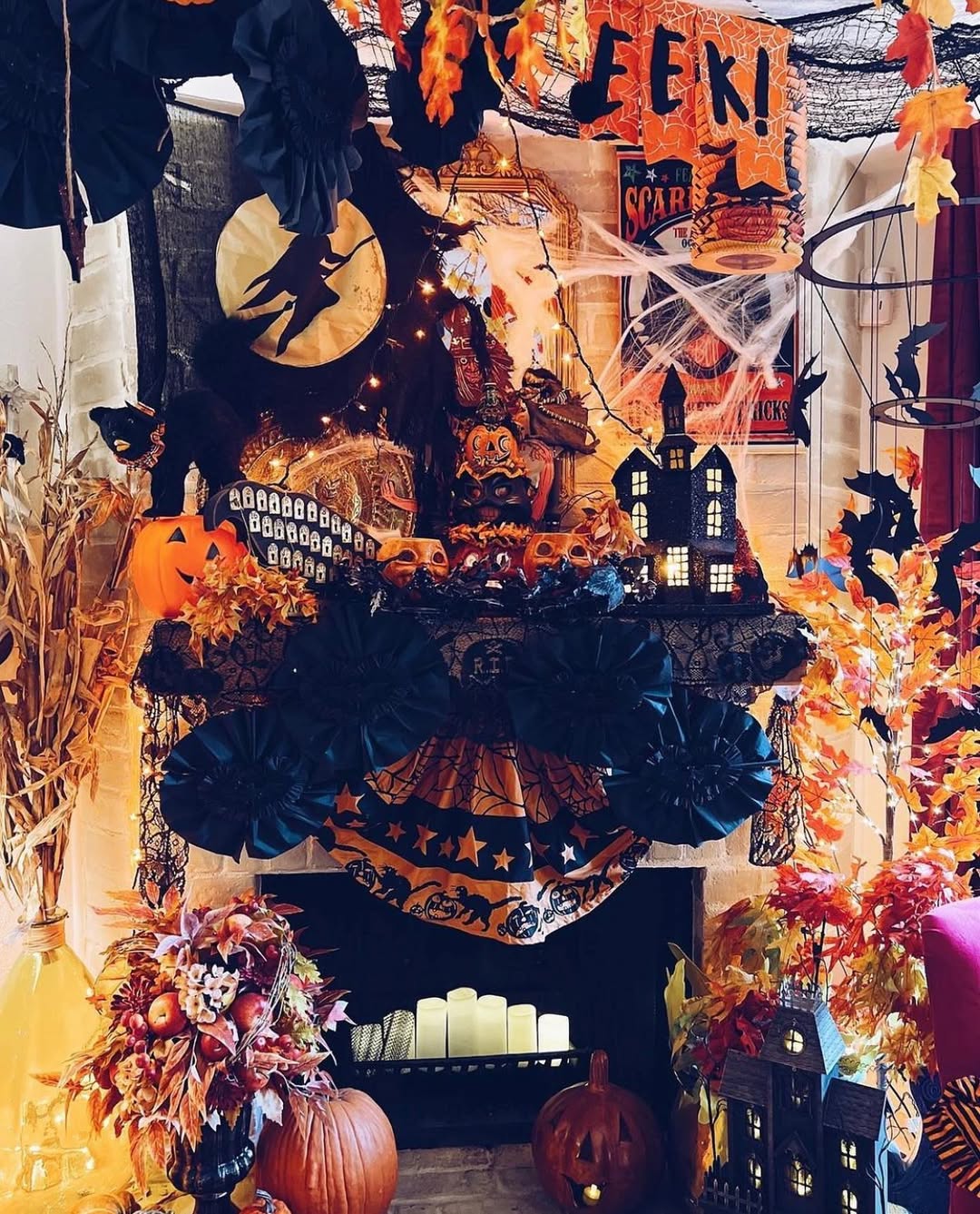A fireplace decorated with Halloween decor, including pumpkins, candles, fake cobwebs, black flowers, autumn leaves, vintage signs, and a witch silhouette in front of a full moon—perfect inspiration for your vintage Halloween ideas.