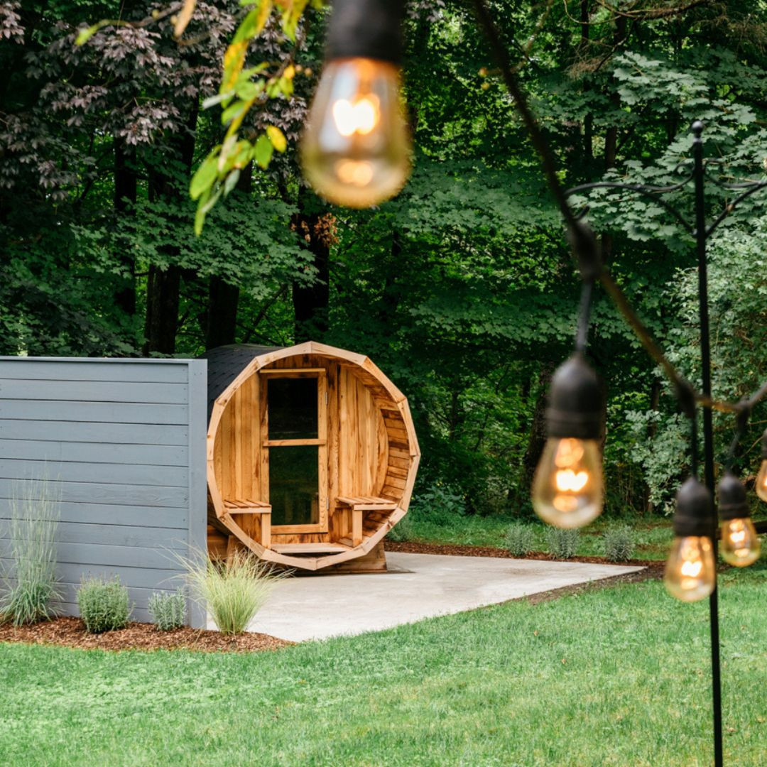 A barrel-shaped wooden sauna sits on a concrete pad beside a gray fence, surrounded by grass, plants, and trees, with string lights in the foreground—perfect inspiration for gorgeous outdoor saunas and stylish sauna design.
