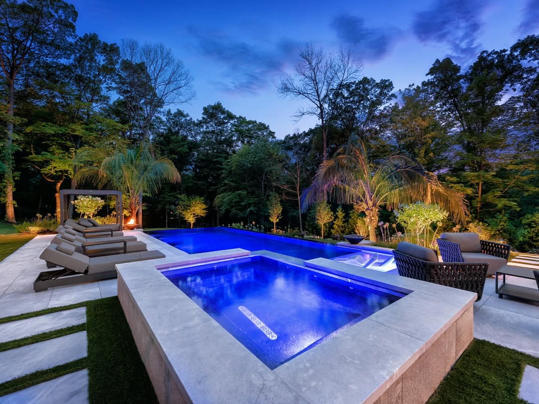 Modern outdoor pool and hot tub area illuminated at dusk, showcasing lounge chairs, palm trees, and lush greenery—perfect inspiration for backyard pool ideas.